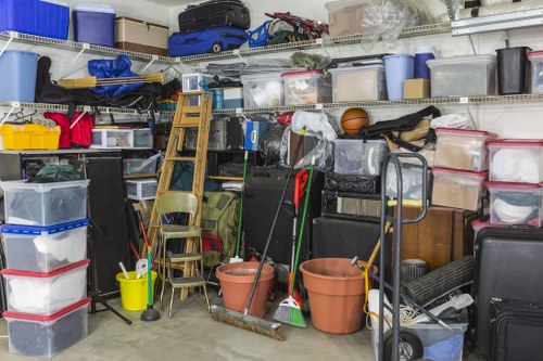 A cluttered storage area within a garage or workshop space containing various plastic storage boxes and crates stacked on metal shelving, some transparent with visible contents such as tools and household items, alongside several gardening buckets, brooms, mops, and a dustpan placed on the floor in front. There are wooden chairs stacked against the shelves and a small step ladder leaning in the background. The environment appears enclosed with visible concrete or painted walls, and natural or artificial lighting creating minimal shadows. The scene depicts an organized but densely packed area suitable for waste collection or rubbish removal, with businesses like Business Waste Removal Tooting specialising in clearing such clutter in the local area near South West London.