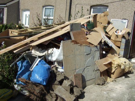A large pile of mixed rubbish accumulated on a pavement outside a residential building, comprising broken wooden planks, cardboard boxes, plastic bags, foam insulation, and construction debris. The wooden planks are light-colored with a rough texture, some leaning against the other waste materials. The cardboard boxes vary in size and shape, with some flattened and others still intact, showing printed markings and torn edges. Blue and white plastic bags are visible, filled with waste, with some spilling contents onto the ground. Concrete blocks and rubble are also present at the base of the pile, exhibiting a rough, coarse texture and grey coloration. In the background, the facades of a brick or pebble-dashed residential building with closed white-window frames can be seen, indicating a typical urban setting, with small plants or shrubs near the building's foundation. The overall scene appears to depict an unmanaged or recently collected rubbish accumulation, consistent with waste removal services provided by Business Waste Removal Tooting in the local community.