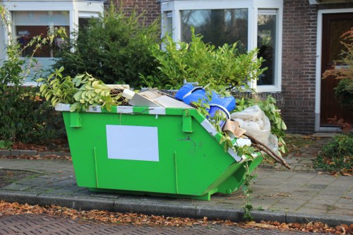 A large green mobile rubbish skip filled with various types of waste materials, positioned on a paved pavement outside a residential building typical of the Tooting area. The skip contains a mix of garden waste including leafy branches and green foliage, along with discarded packaging, cardboard boxes, plastic items, and fabric debris. It is situated adjacent to a curb, with a concrete border separating it from the road, and the background shows the front facade of a brick house with white-framed windows and a small garden area with shrubs. The scene is lit by natural daylight, highlighting the contrasting textures and colors of the waste and the surrounding environment. This image relates to rubbish removal services provided by Business Waste Removal Tooting, supporting waste collection from residential or commercial properties in the area. The overall composition emphasizes the importance of efficient waste management and clearance within the local community.