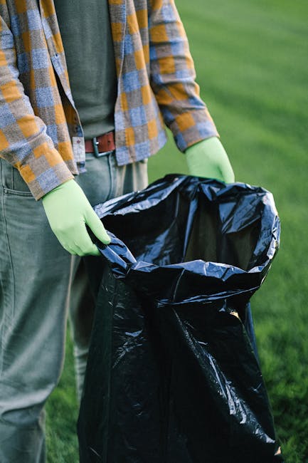 A person wearing a plaid shirt with shades of yellow, gray, and blue, a green long-sleeved layer underneath, and gray pants secured with a brown belt, is standing outdoors on a grassy area. They are h