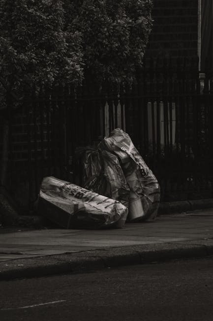 A roadside scene featuring two large black plastic rubbish bags filled with waste material, positioned on the pavement close to a dark wooden fence and a dense leafy hedge in the background. The bags 